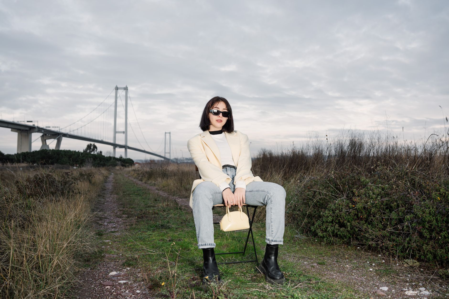 Stylish woman in sunglasses sitting by a bridge, showcasing modern fashion against a natural backdrop. — Need Help Fast? How